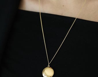 May include: A gold-colored locket necklace with a round pendant. The pendant hangs from a delicate gold chain. The necklace is displayed against a black background.