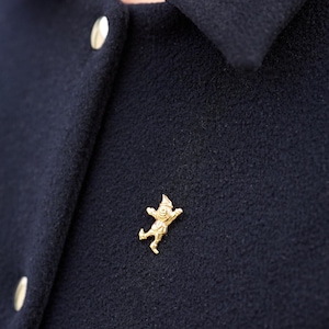 Puede incluir: Un broche dorado con forma de figura caprichosa, sujeto a una chaqueta azul marino oscuro. El broche presenta un personaje sonriente con un sombrero puntiagudo y los brazos extendidos. La chaqueta tiene botones dorados.