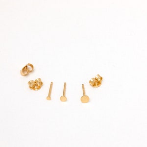 May include: Three pairs of gold stud earrings with different sized round discs. The earrings are on a white background.