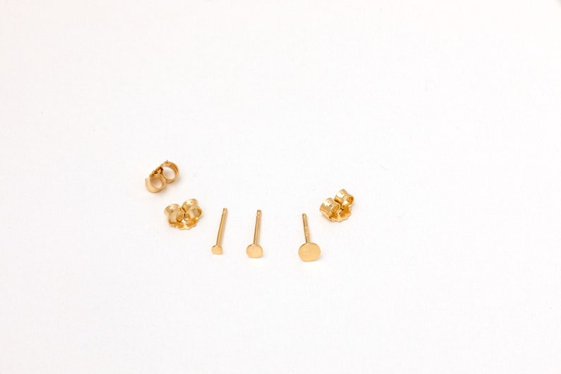 May include: Three pairs of gold stud earrings with different sized round discs. The earrings are on a white background.