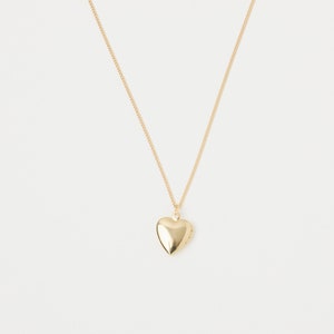 Op de afbeelding: Een goudkleurige hartvormige medaillon ketting. Het medaillon hangt aan een delicate ketting tegen een witte achtergrond. De hartvormige hanger is glad en reflecterend, wat wijst op een gepolijste afwerking.
