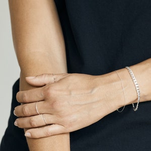 May include: A person's arm wearing a silver chain bracelet and two thin silver rings on their fingers.