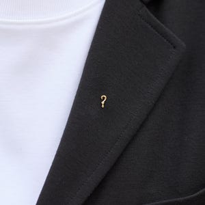 Könnte beinhalten: Ein goldfarbener Fragezeichen-Ansteckpin auf einem schwarzen Blazer. Der Pin ist klein und schlicht und verleiht eine subtile Note. Ein weißes Rundhals-T-Shirt ist darunter sichtbar.