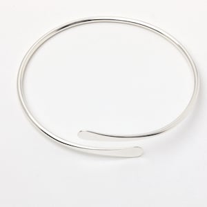 Op de afbeelding: Een zilveren armband met een eenvoudig, open ontwerp. De armband is gemaakt van een enkel, dun stuk metaal dat in een cirkel is gebogen. De uiteinden van de armband zijn met een klein, plat stuk metaal aan elkaar verbonden.