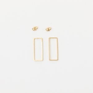 Könnte beinhalten: Ein Paar goldfarbene rechteckige Ohrstecker und zwei Ohrringverschlüsse auf weißem Hintergrund. Die Ohrringe haben ein einfaches, offenes Rahmendesign. Die Ohrringverschlüsse sind klein und strukturiert.