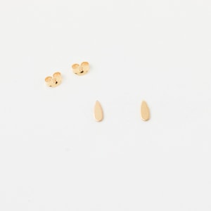 May include: A pair of gold-colored teardrop earrings and two butterfly earring backs are displayed on a white surface. The earrings have a smooth, polished finish and a simple, elegant design. The earring backs are also gold-colored.