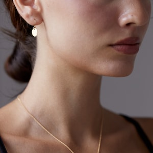Op de afbeelding: Een gouden ketting met een klein rond hangertje. De ketting wordt gedragen door een persoon met bruin haar en een zwart shirt.