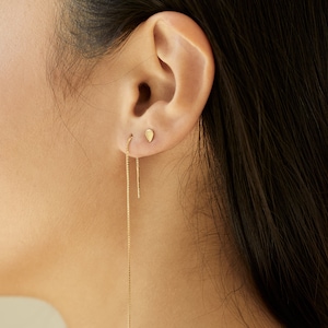 Op de afbeelding: Een gouden kettingoorring met een kleine traanvormige oorbel. De oorring wordt gedragen in het linker oor van een persoon met lang zwart haar.