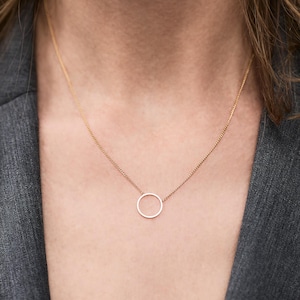 Könnte beinhalten: Eine zarte, goldfarbene Halskette mit einem einfachen, offenen Kreisanhänger. Die Halskette wird über einer grauen Jacke getragen, was eine minimalistische und elegante Ästhetik erzeugt. Die Kette ist fein und der Anhänger ist zentriert.