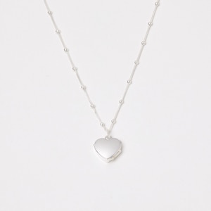 Op de afbeelding: Een zilveren ketting met een hartvormige medaillonhanger. De ketting heeft kleine, gelijkmatig verdeelde kralen. Het medaillon is gesloten en glad, waardoor het licht wordt gereflecteerd. Op een witte achtergrond.