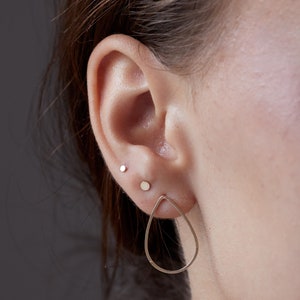 May include: A person wearing a pair of gold teardrop earrings. The earrings are simple and delicate, with a smooth, polished finish.