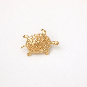 Könnte beinhalten: Eine goldfarbene Schildkrötenbrosche. Die Brosche zeigt ein detailliertes Design einer Schildkröte mit strukturiertem Panzer, vier Beinen, einem Kopf und einem kleinen Schwanz. Die Schildkröte ist vor einem weißen Hintergrund platziert.