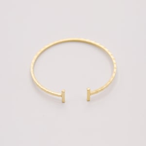 Könnte beinhalten: Ein goldfarbenes, offenes Armreif-Armband mit gehämmertem Muster. Das Armband hat ein schlichtes Design mit zwei rechteckigen Endstücken. Der Schmuck wird auf weißem Hintergrund präsentiert.