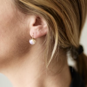 May include: A gold-colored seashell-shaped earring is worn on the ear. The earring has a textured, fan-like design. The image shows the side of a person's head with blonde hair and a black top.