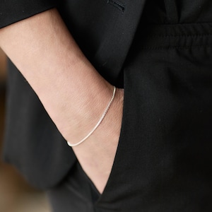May include: A delicate silver chain bracelet. The bracelet is worn on the wrist, visible against the dark fabric of a black jacket. The chain is thin and appears to be made of small, interconnected links.