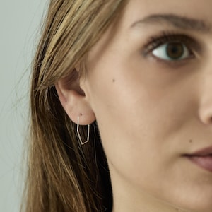 Puede incluir: Primer plano de una oreja con un pendiente geométrico plateado. El pendiente presenta un diseño angular único en forma de pentágono. Fabricado en metal fino y brillante, muestra un estilo minimalista y moderno.