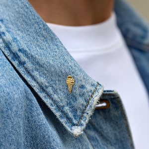 Könnte beinhalten: Eine goldfarbene Eiswaffel-Anstecknadel ist am Revers einer hellblauen Jeansjacke befestigt. Die Jacke wird über einem weißen T-Shirt getragen. Die Anstecknadel verleiht dem lässigen Outfit einen Hauch von Verspieltheit.