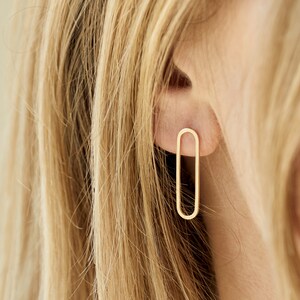 Könnte beinhalten: Goldfarbene, minimalistische ovale Ohrringe. Die Ohrringe haben eine einfache, längliche ovale Form mit glatter, polierter Oberfläche. Die Ohrringe werden am Ohr einer Person gezeigt, mit blondem Haar im Hintergrund.