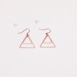 Op de afbeelding: Een paar roségoudkleurige bungelende oorbellen met een geometrisch driehoeksontwerp. Elke oorbel bestaat uit een groter buitenste driehoekig frame met een kleinere horizontale balk in het midden, hangend aan een gebogen oorhaakje met doorzichtige sluitingen.