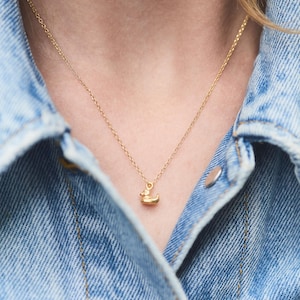 Könnte beinhalten: Eine goldene Kettenkette mit einem kleinen, goldenen Entenanhänger. Die Kette wird von einer Person getragen, die eine Jeansjacke trägt.