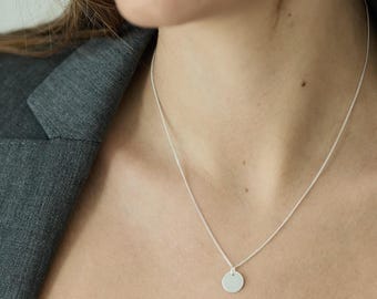 May include: A silver necklace with a small, round pendant. The pendant hangs from a delicate chain. The necklace is worn against a gray blazer.