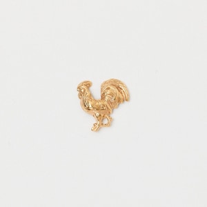 Puede incluir: Figura de gallo dorada. El gallo está de pie con la cabeza en alto y las plumas de la cola extendidas. La figura es muy detallada, con rasgos intrincados. El gallo es un símbolo de coraje y orgullo.