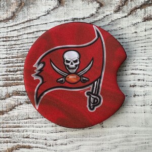 May include: A red, white, and black round coaster with the Tampa Bay Buccaneers logo. The logo features a skull and crossbones with a football in the center.