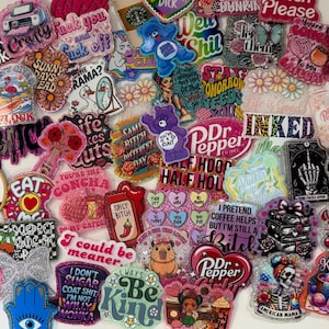 May include: A collection of colorful, glittery acrylic pins and patches with various designs and text. The pins feature images of bears, flowers, food, and phrases like "Be Kind" and "I Pretend Coffee Helps".