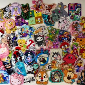 May include: A collection of colorful, cartoon-themed charms featuring characters from various animated shows and movies. The charms include characters like Hello Kitty, Deadpool, and Disney characters. The charms are made of a shiny, possibly acrylic material.