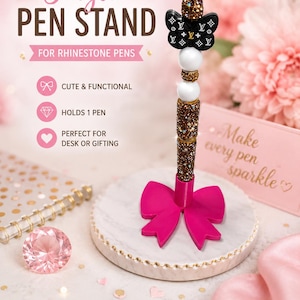 May include: A single pen stand featuring a circular white marble-patterned base with a bright pink bow accent. The vertical holder is decorated with stacked beads, including a black butterfly charm with white logos and brown glitter accents.