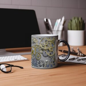 May include: A black and white color changing mug with a design of gold and silver gears. The mug is on a wooden table with a computer, keyboard, and other office supplies.