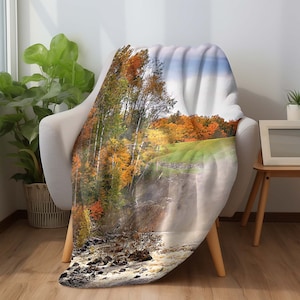 May include: A soft, plush throw blanket featuring a scenic autumn landscape with a waterfall. The blanket showcases vibrant fall foliage in shades of red, orange, and yellow, with a cascading waterfall in the foreground.