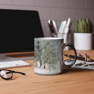 May include: A black and white magic mug with a snowy forest scene. The mug is on a wooden table with a keyboard, glasses, and a pen.