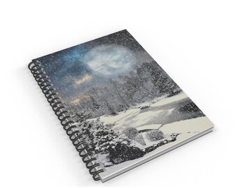 May include: A spiral bound notebook with a winter scene on the cover. The cover features a snowy forest with a large, full moon in the sky. The moon is surrounded by a blue and purple sky with white clouds. The forest is covered in snow and there is a frozen lake in the foreground.