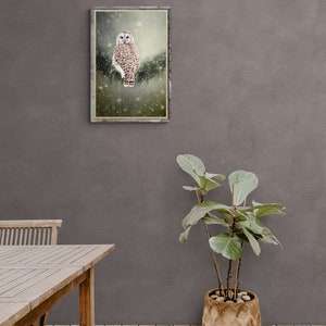 May include: A framed print of a brown and white owl perched on a branch in a snowy forest. The owl has large, yellow eyes and is looking directly at the camera. The print is hanging on a gray wall above a wooden table and a potted plant.