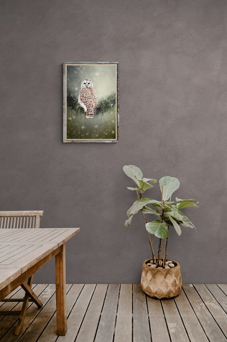 May include: A framed print of a brown and white owl perched on a branch in a snowy forest. The owl has large, yellow eyes and is looking directly at the camera. The print is hanging on a gray wall above a wooden table and a potted plant.