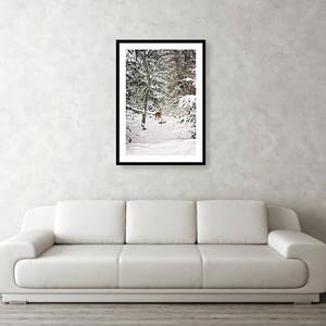 K&ouml;nnte beinhalten: Gerahmter Kunstdruck einer winterlichen Waldszene. Das Kunstwerk zeigt einen schneebedeckten Wald mit B&auml;umen und einem Hirsch. Der Druck ist in einem schwarzen Rahmen und wird &uuml;ber einem wei&szlig;en Sofa vor einer neutralen Wand ausgestellt.