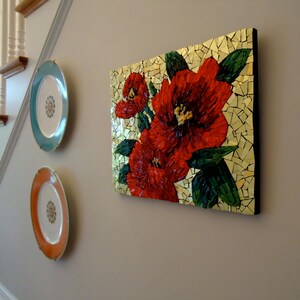 K&ouml;nnte beinhalten: Ein Mosaik-Blumenkunstwerk mit leuchtend roten Blumen mit gr&uuml;nen Bl&auml;ttern vor goldenem Hintergrund. Zwei dekorative Teller mit floralen Designs in T&uuml;rkis und Orange h&auml;ngen an der Wand und erg&auml;nzen die Farben des Kunstwerks.