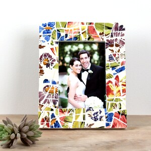 May include: A rectangular photo frame with a mosaic tile design. The frame features a colorful floral pattern with blue, red, yellow, and purple hues. The frame holds a photograph of a smiling couple. The frame is on a wooden surface.