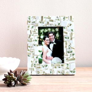 May include: A rectangular photo frame with a mosaic design. The frame is made of white tiles with green and gold accents. The frame holds a photo of a smiling couple. The frame is on a wooden surface.