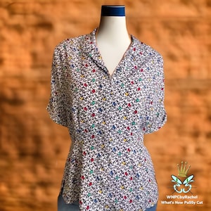 May include: A white blouse with a v-neck and short sleeves. The blouse has a small, colorful floral print.