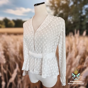 May include: White lace blouse with a V-neckline, long sleeves, and a fitted waist. The blouse has a delicate floral pattern and a satin ribbon tie at the waist.