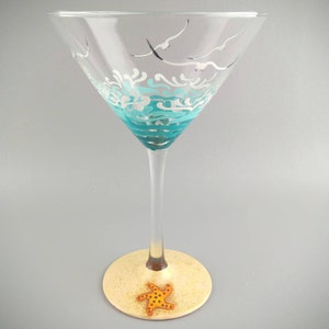 May include: A turquoise and white hand-painted martini glass with a sea-themed design. The base of the glass is painted to look like sand with a starfish.