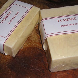 May include: Two bars of turmeric goat's milk soap, wrapped in clear plastic and labeled. The labels read "TUMERIC GOATS MILK SOAP" in a serif font with a red border. The soap has a light yellow color.