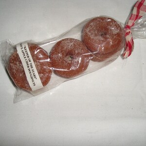 May include: Three apple cider donuts in a clear plastic bag tied with a red and white checkered ribbon. The bag has a label that reads "Keeper of the Stars" and "Apple Cider Doughnuts".