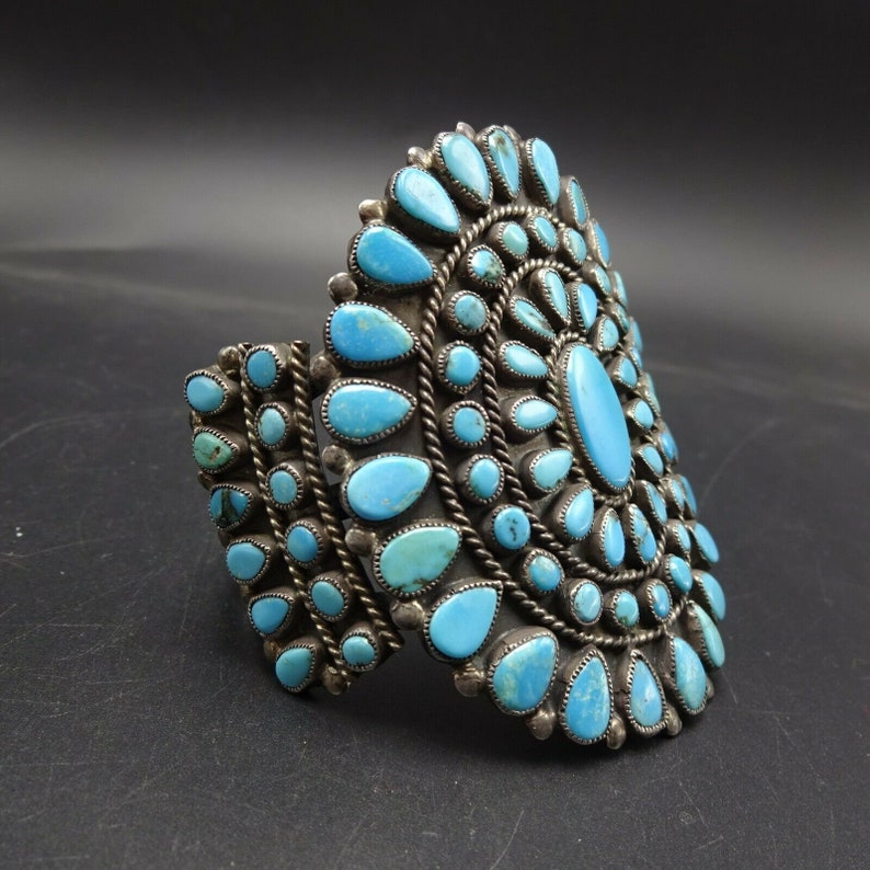 May include: A silver cuff bracelet with a large turquoise stone cluster in the center. The turquoise stones are set in a circular pattern and are surrounded by smaller turquoise stones.