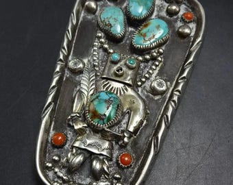 Puede incluir: Corbata bolo plateada con un colgante rectangular adornado con turquesas y coral. El colgante presenta una figura central con una piedra turquesa, rodeada de piedras turquesas, cuentas de coral y detalles plateados decorativos. Cordón de cuero negro.