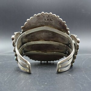 May include: A silver and turquoise bracelet with a decorative design. The bracelet has three bands and is stamped with the initials "V.M.B."