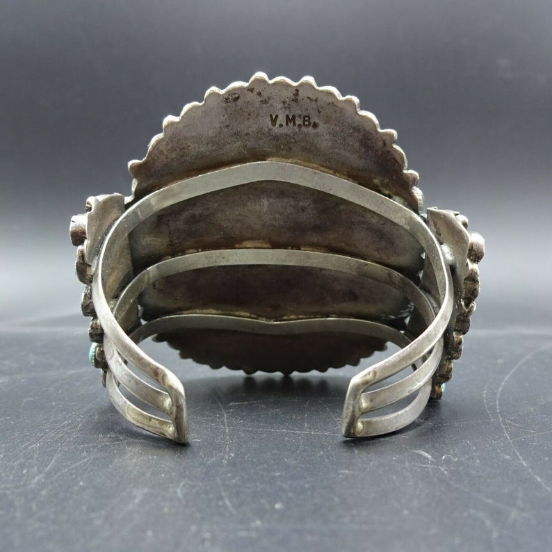 May include: A silver and turquoise bracelet with a decorative design. The bracelet has three bands and is stamped with the initials "V.M.B."