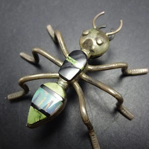 May include: A silver-toned metal insect brooch featuring an elongated body with inlaid segments. The thorax contains a black stone with a green vein, while the abdomen displays a triangular iridescent opal-like stone and a green stone, separated by black bands.